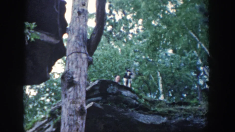 1964 Wisconsin home-movie frame showing two distant figures on a rocky ledge among trees, partly obscured by foliage and soft focus.