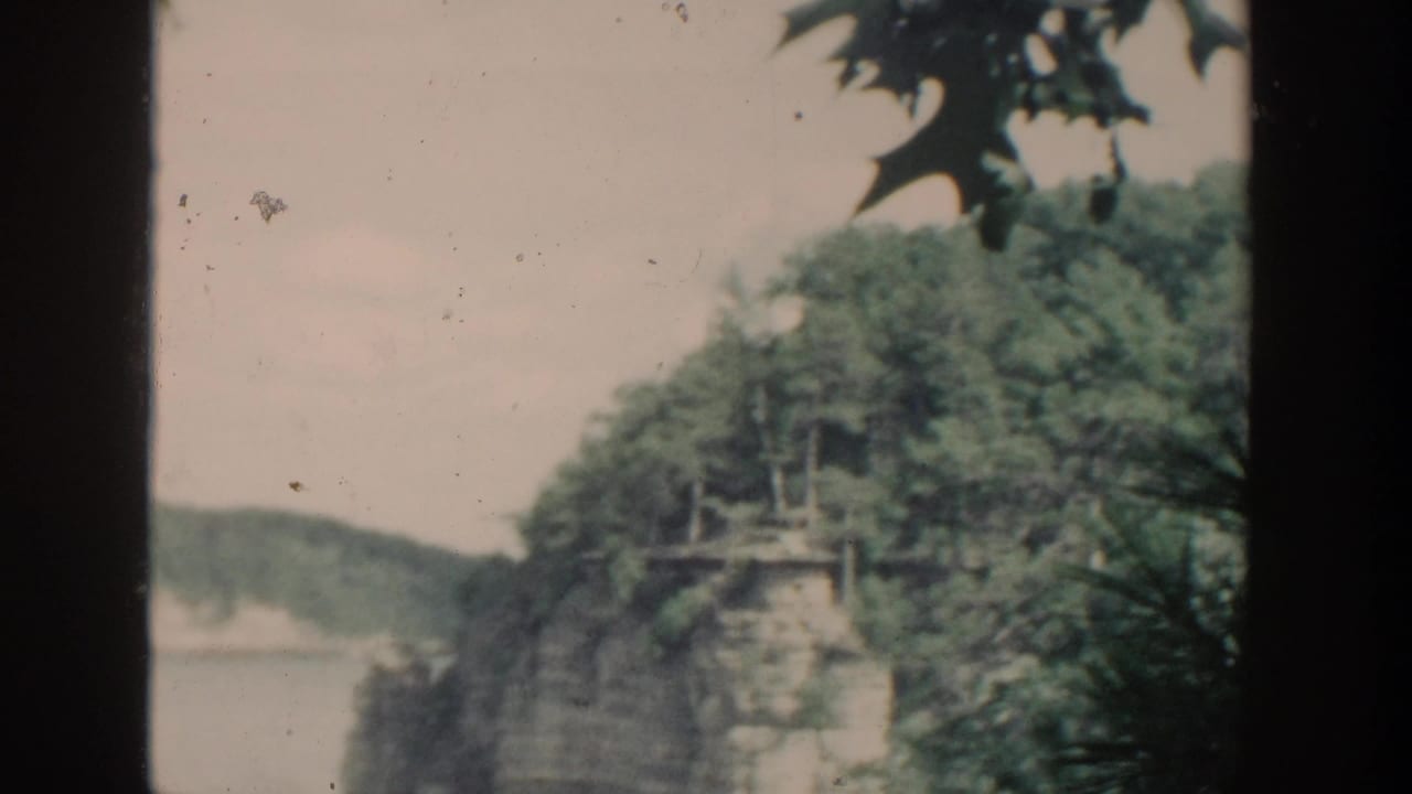 1964 Wisconsin 8mm frame showing calm water, tree-covered rock cliffs, dark window-like edges, and foreground foliage.