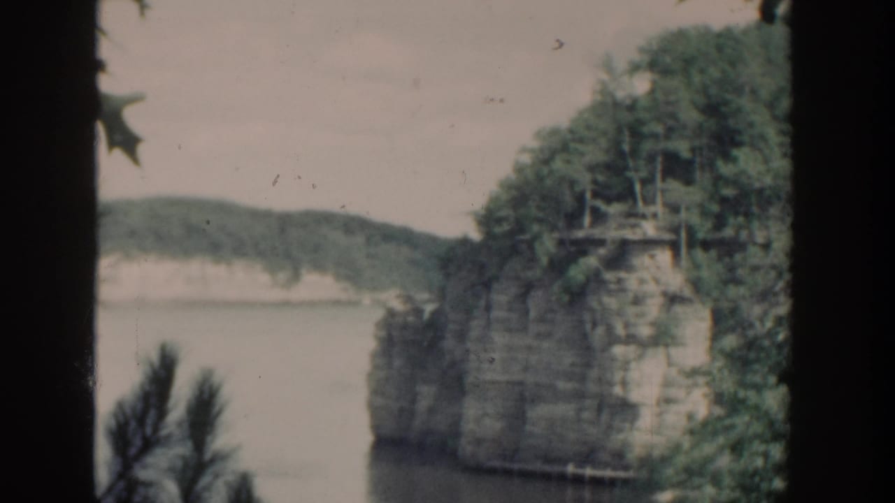 Rocky shoreline cliff with trees above calm water, partially framed by a dark vertical edge.