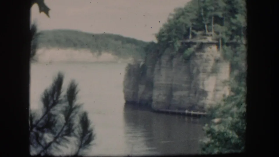 1964 Wisconsin 8mm still with calm lake water, wooded rock cliffs, a foreground pine branch, and soft distant hills.