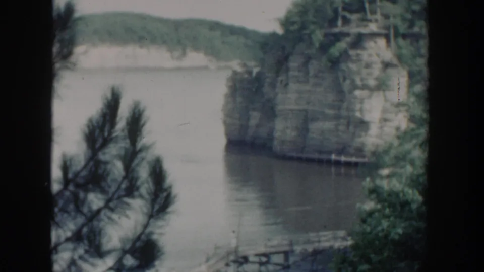1964 Wisconsin frame showing a wooded rocky cliff at the water's edge, foreground pine needles, and a hazy lake backdrop.