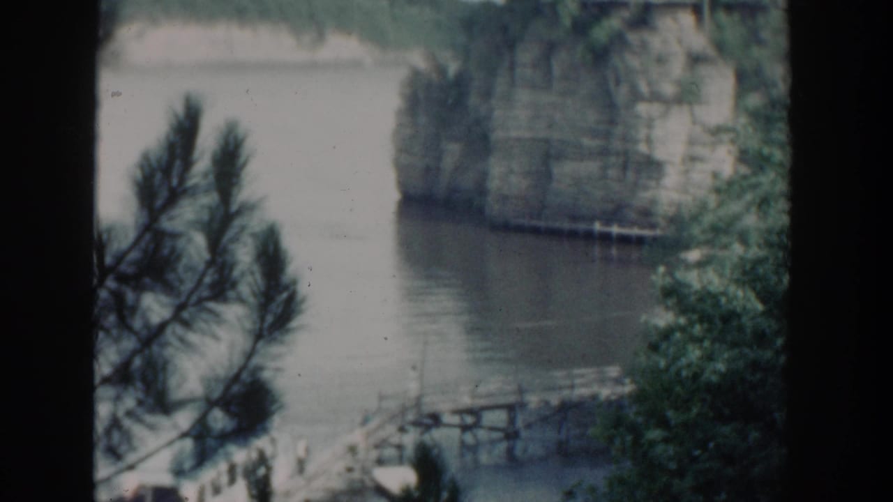 1964 Wisconsin 8mm frame showing pine branches over calm water, a small docked white boat, a wooden pier, and a sheer rock face across the lake.