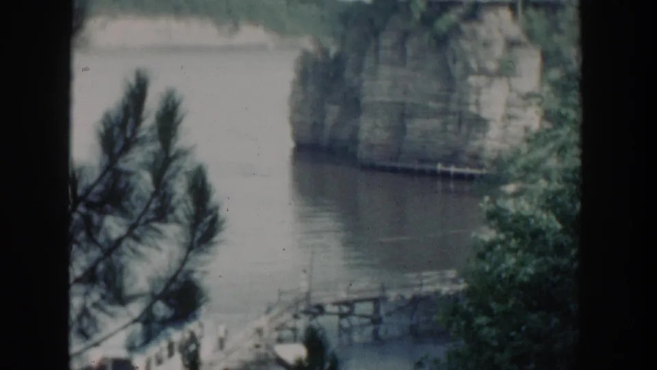 1964 Wisconsin 8mm frame showing pine branches over calm water, a small docked white boat, a wooden pier, and a sheer rock face across the lake.