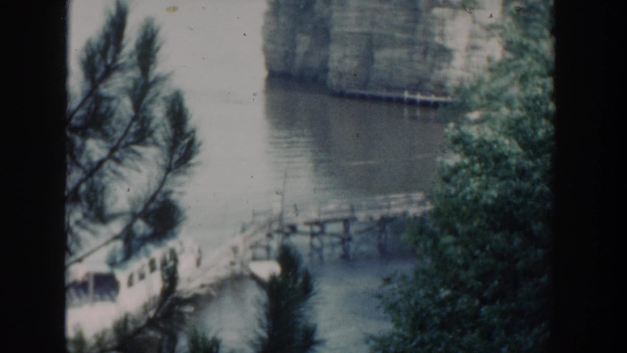 1964 Wisconsin lakeside view with a white boat at a wooden pier, foreground pine needles, still water, shoreline trees, and rock wall beyond.