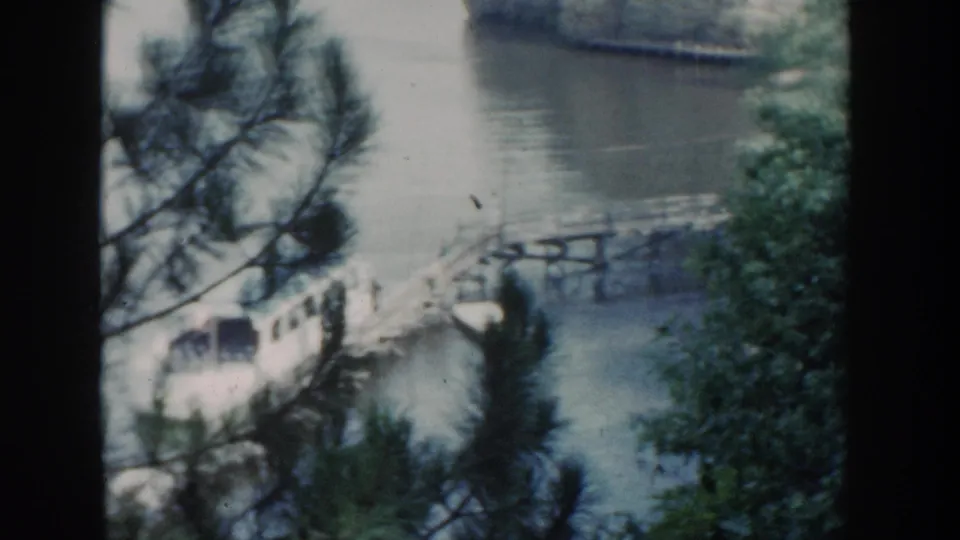 Soft 8mm frame from 1964 Wisconsin showing pine limbs in front of a lakeside pier, a white boat, calm water, leafy trees, and a rock formation.