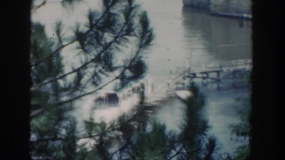 1964 Wisconsin 8mm frame with pine branches in the foreground, a white boat near a wooden pier, calm water, trees, and a pale rock formation.
