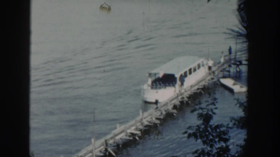 1964 Wisconsin 8mm frame showing a white passenger boat beside a wooden pier on a calm lake, partly obscured by film-frame shadows.
