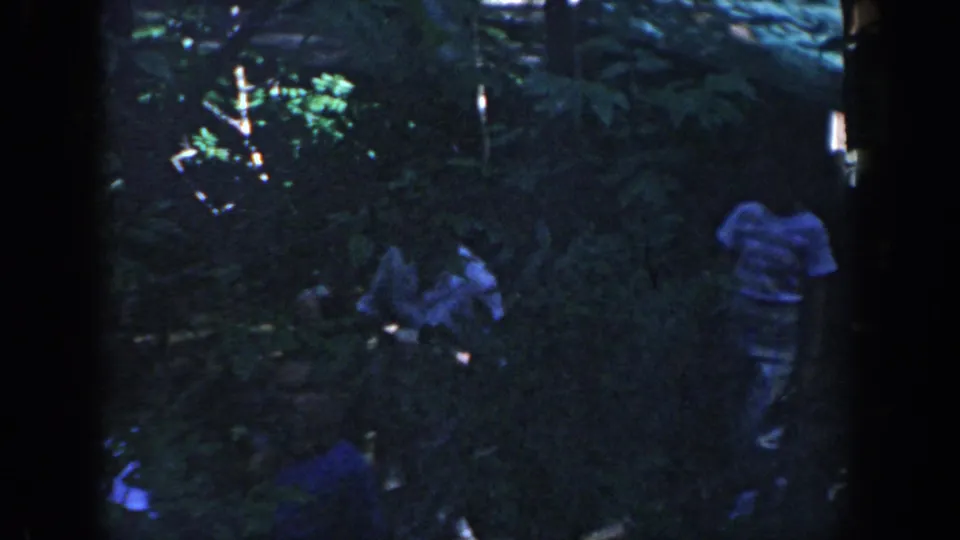 1964 Wisconsin home-movie frame showing several people on a shaded forest trail, partly obscured by dense plants and dark frame edges.