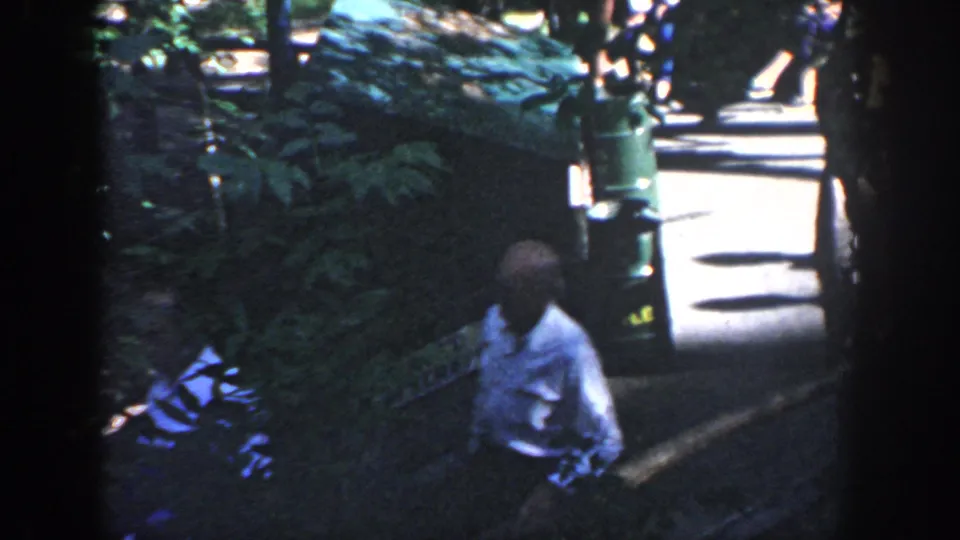 Wisconsin 1964 frame of a man near a bench, green trash can, shaded planting, and bright walkway in a tourist setting.