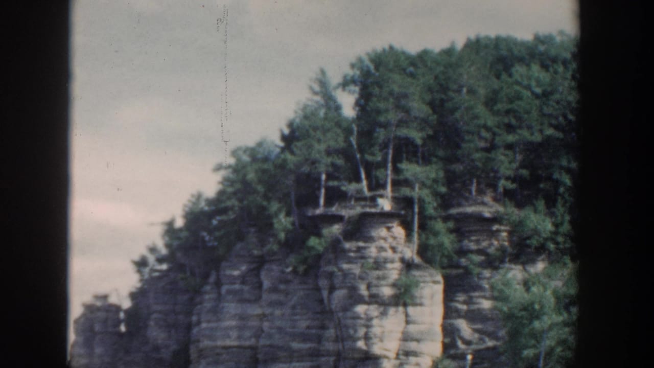 1964 Wisconsin 8mm frame showing a wooded layered rock cliff rising from calm water, seen from a boat with dark film borders.