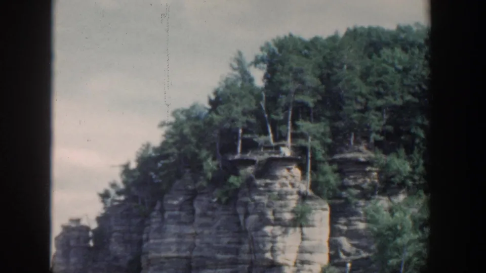 1964 Wisconsin 8mm frame showing a wooded layered rock cliff rising from calm water, seen from a boat with dark film borders.