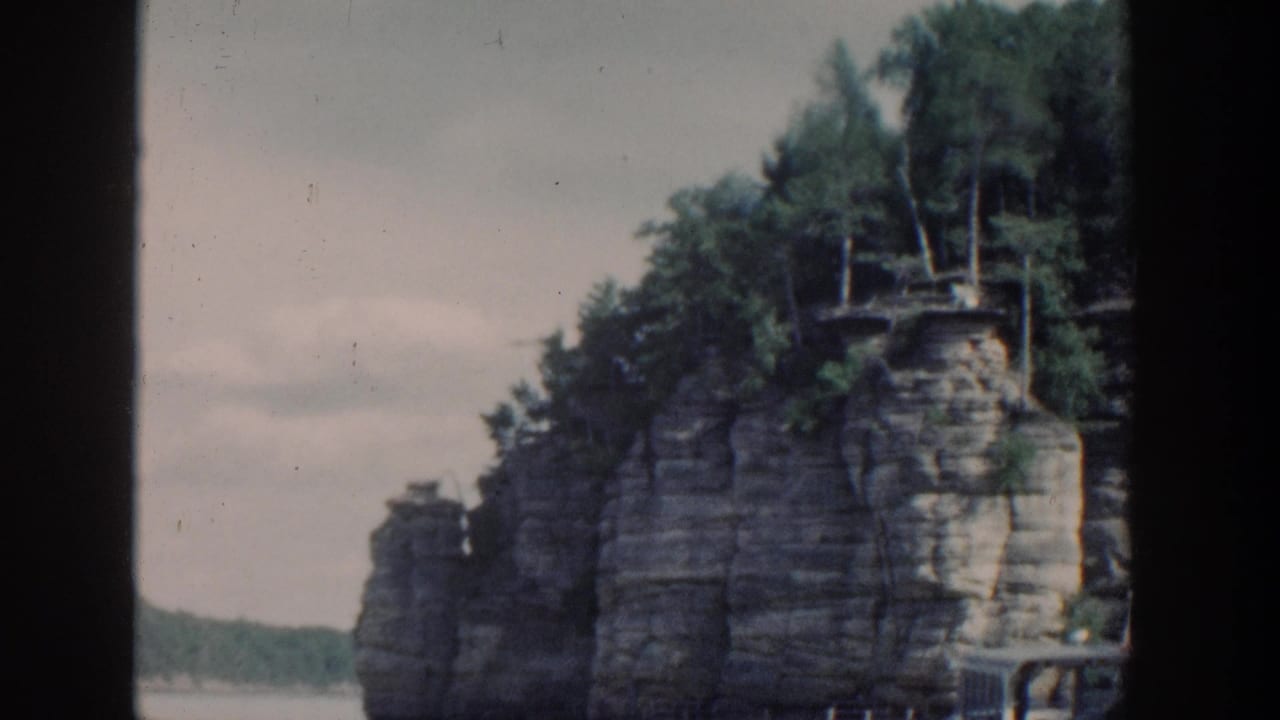 Tall rock formation at the water edge with trees above and a railing near the lower right.