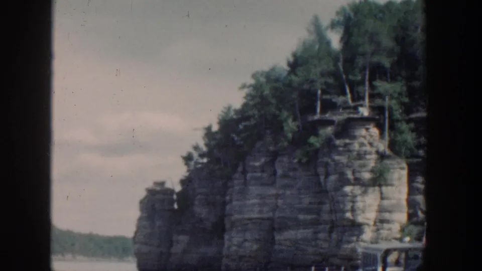 1964 Wisconsin 8mm view of a steep layered cliff beside water, with dense trees above and a small railing or dock edge visible.