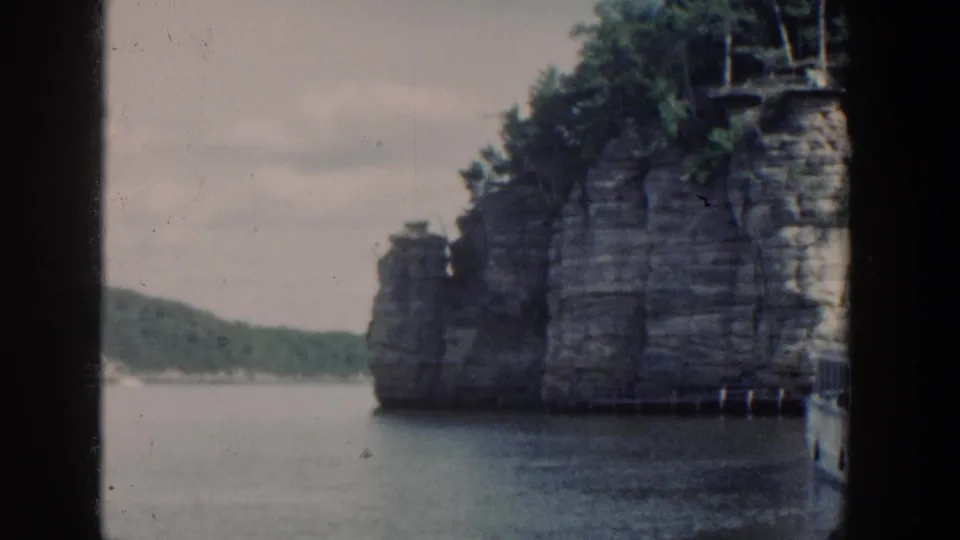 1964 Wisconsin home-movie frame showing a layered cliff at the water edge with forested shoreline and soft 8mm film texture.