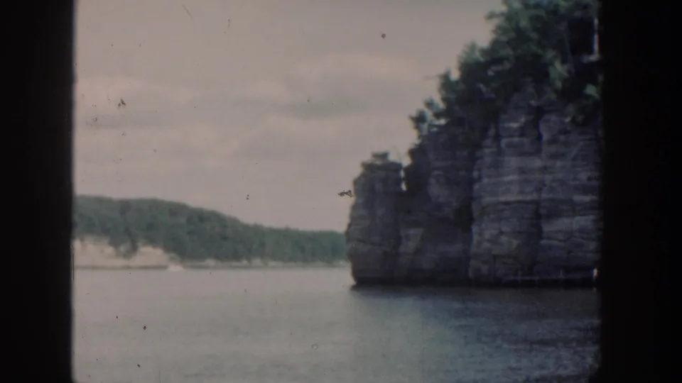 1964 Wisconsin 8mm frame with a wooded rock cliff at right, calm water below, and distant tree-covered shoreline beyond.