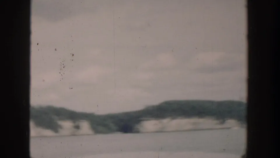 Soft 8mm frame of a Wisconsin lake shoreline with pale cliffs, wooded hills, muted color, and visible film wear.