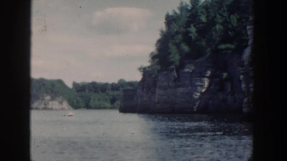 1964 Wisconsin 8mm boat-view frame with a rocky wooded cliff at right, lake water below, and distant shoreline beyond.