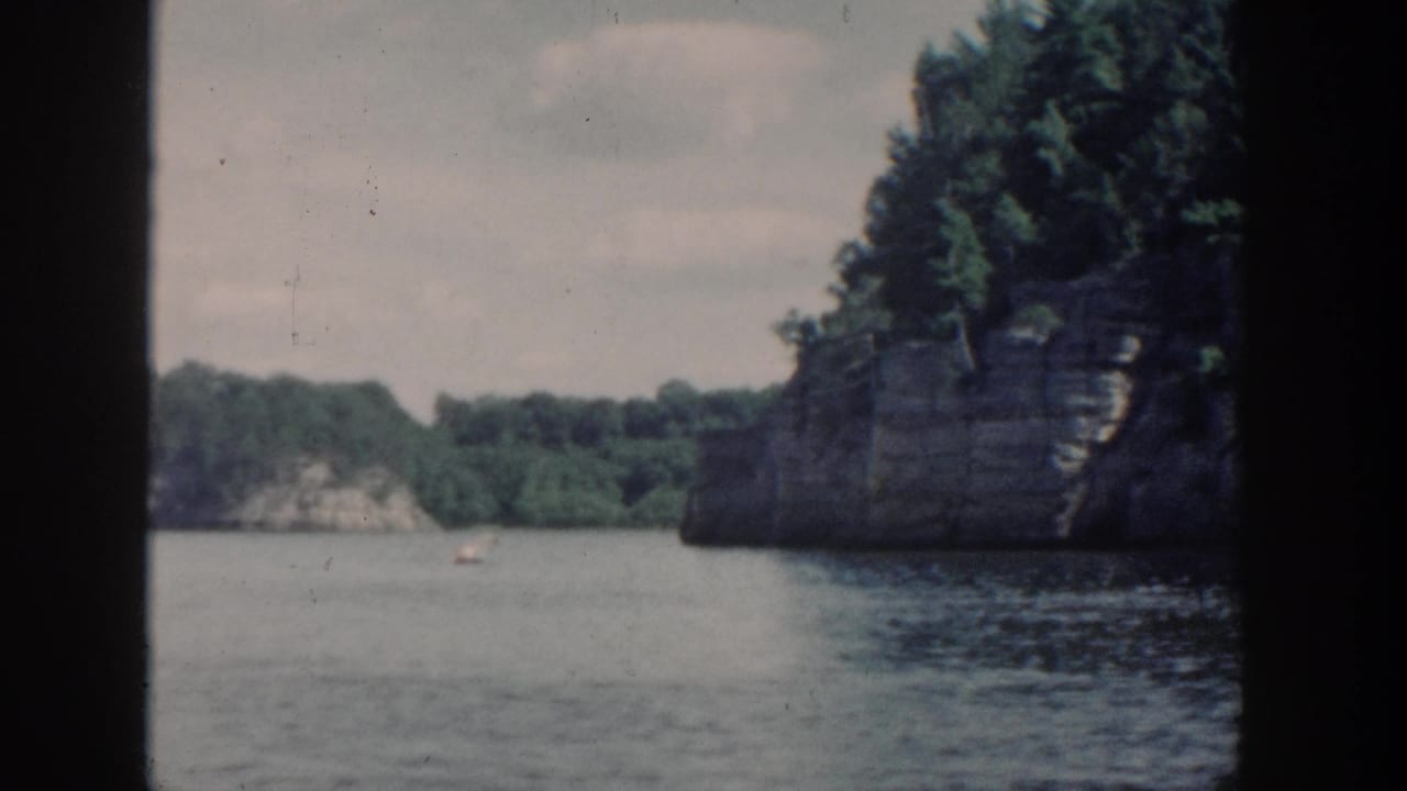 1964 Wisconsin 8mm boat-view frame showing calm lake water, wooded rock formations, and an overcast sky.