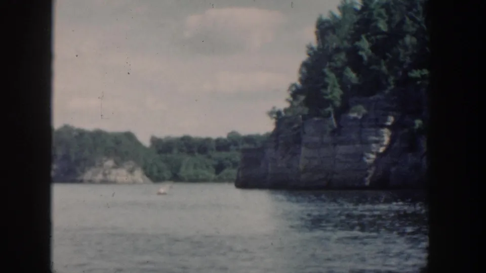 1964 Wisconsin 8mm boat-view frame showing calm lake water, wooded rock formations, and an overcast sky.