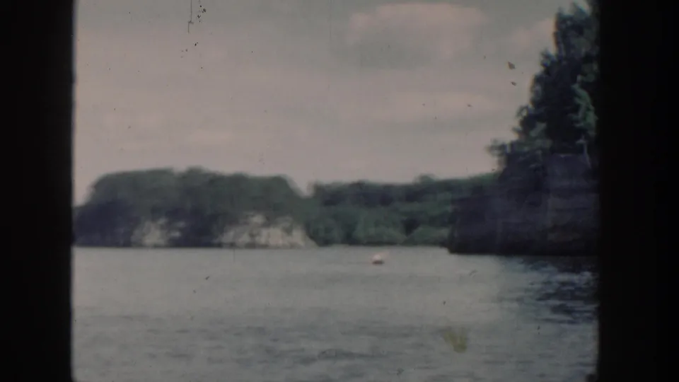 1964 Wisconsin lake view with forested cliffs, muted water surface, and a small distant boat-like shape.