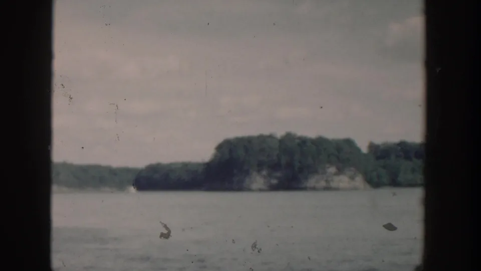 1964 Wisconsin lake scenery with a distant wooded bluff, calm water, cloudy sky, and visible 8mm film wear.
