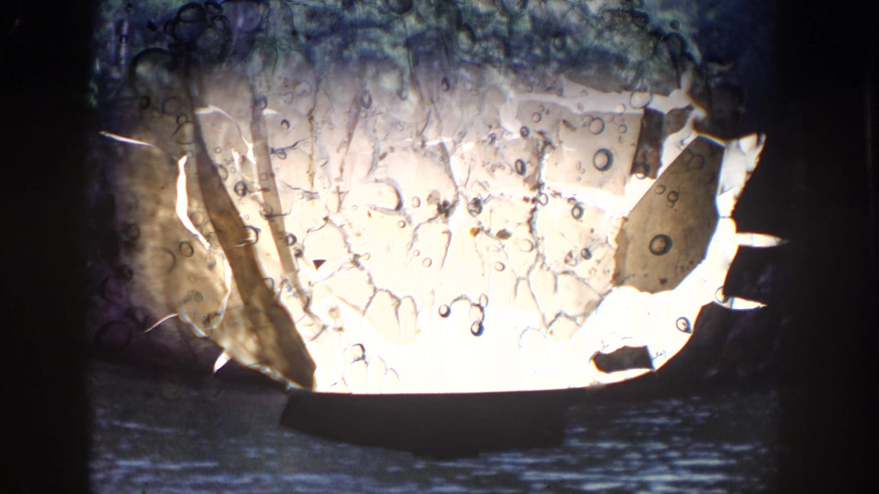 1964 Wisconsin 8mm frame showing film damage over a dark lake view with wooded shoreline and rocky terrain in the background.