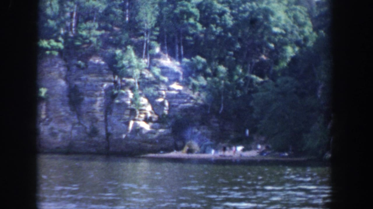 Rocky cliff and dense trees across a lake, with small shoreline figures or campsite details visible.