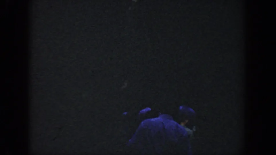 Low-exposure 1964 Wisconsin home-movie frame with shadowed people low in the frame and minimal visible lakeside detail.