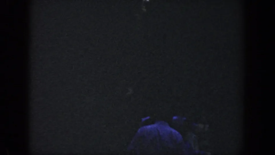 1964 Wisconsin home-movie frame showing a barely visible figure in a light shirt moving through a dark wooded night scene.