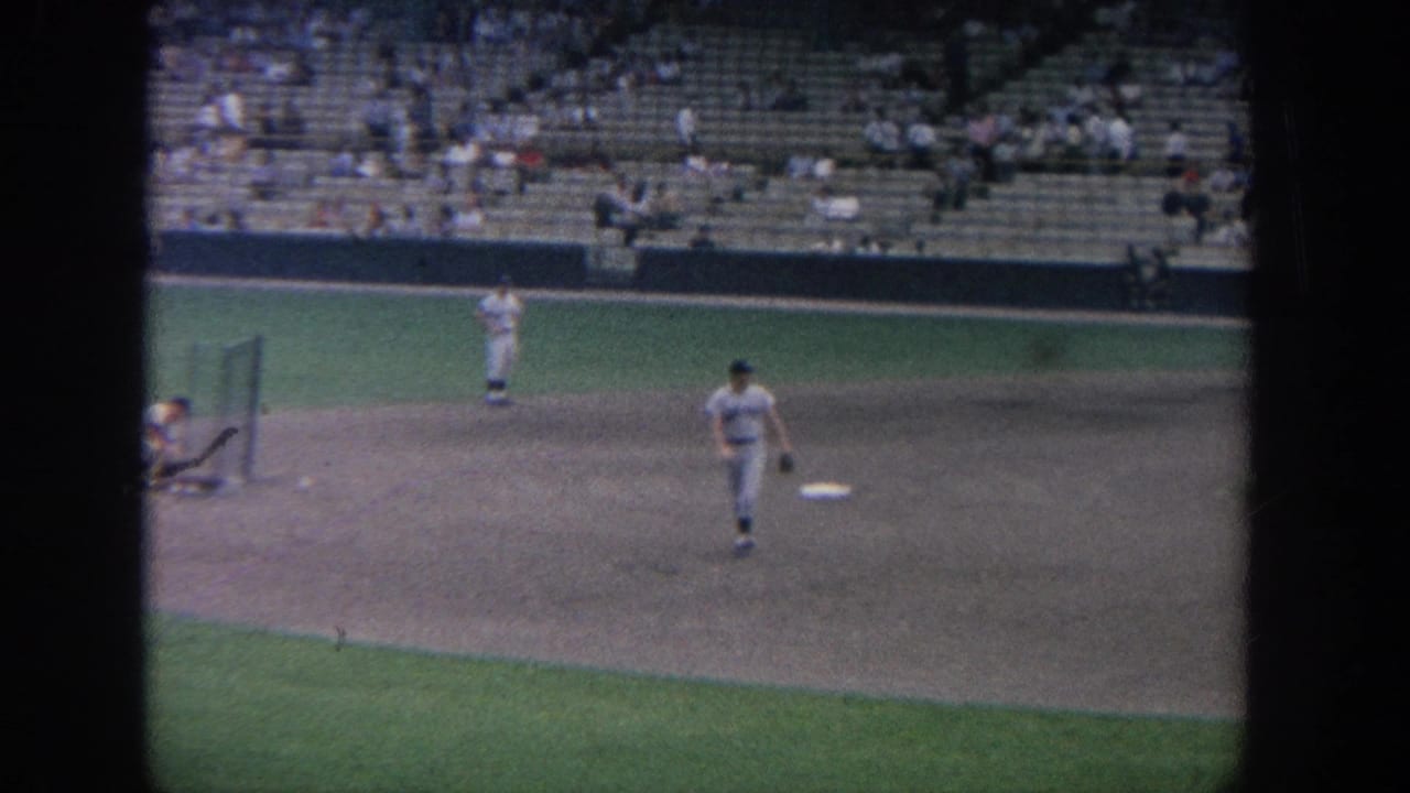 1964 New York 8mm frame showing baseball players spaced across the infield as spectators sit in the stands.
