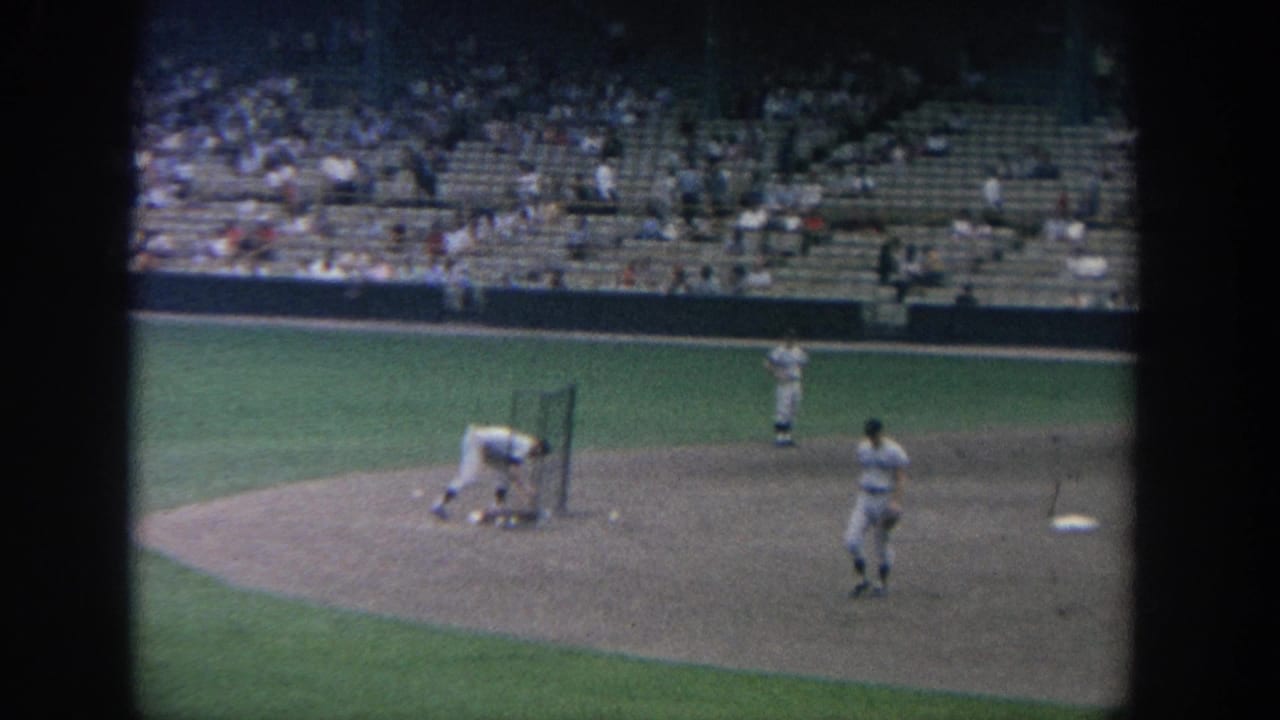 A baseball player bends near a batting screen while others stand on the infield.