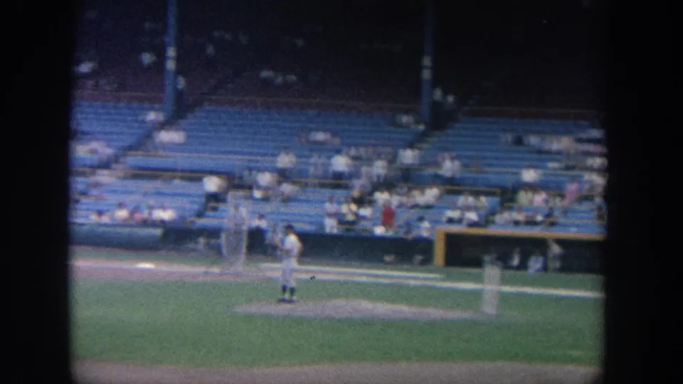 1964 New York stadium frame showing a pitcher near the mound, a field screen, and spectators seated in the background.