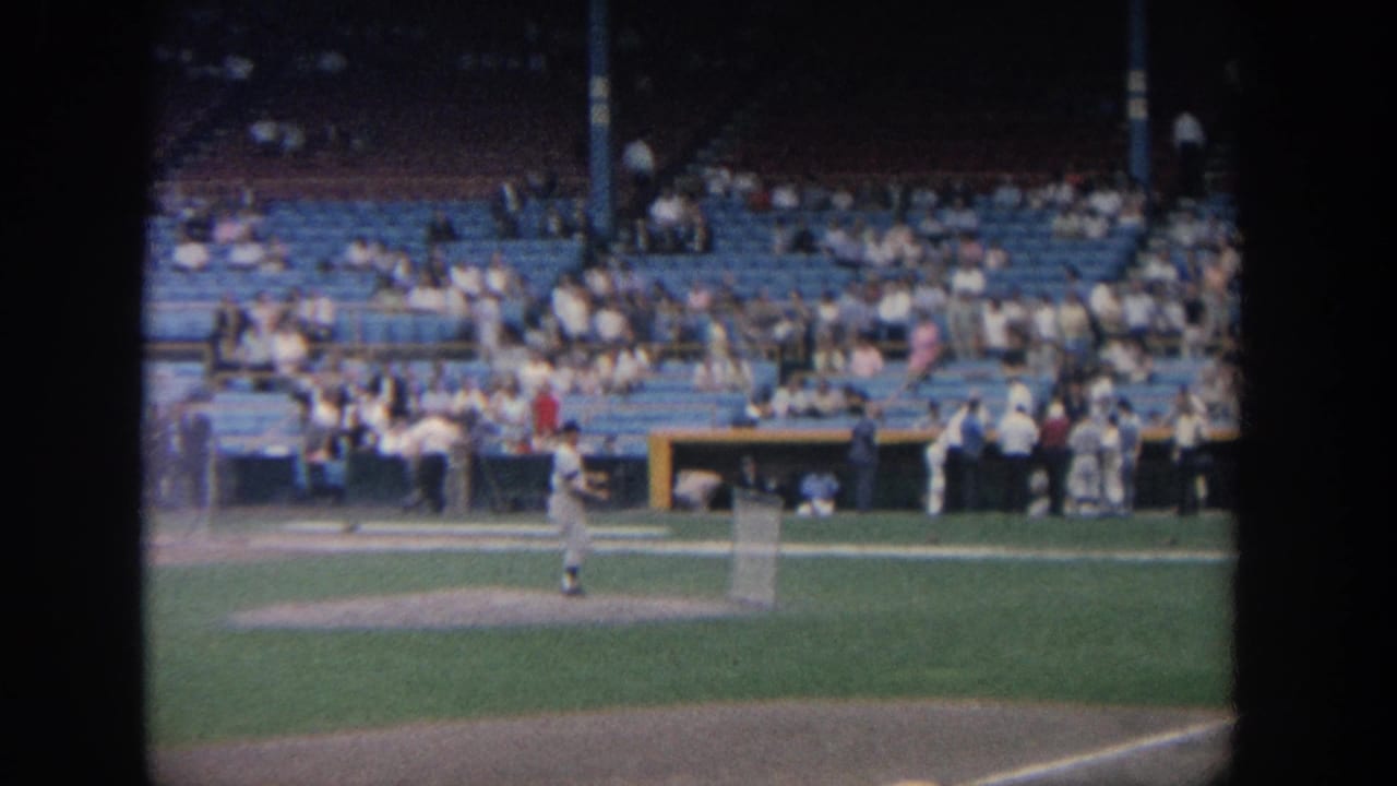 1964 New York 8mm frame showing baseball players warming up near the infield as spectators sit in the stands.