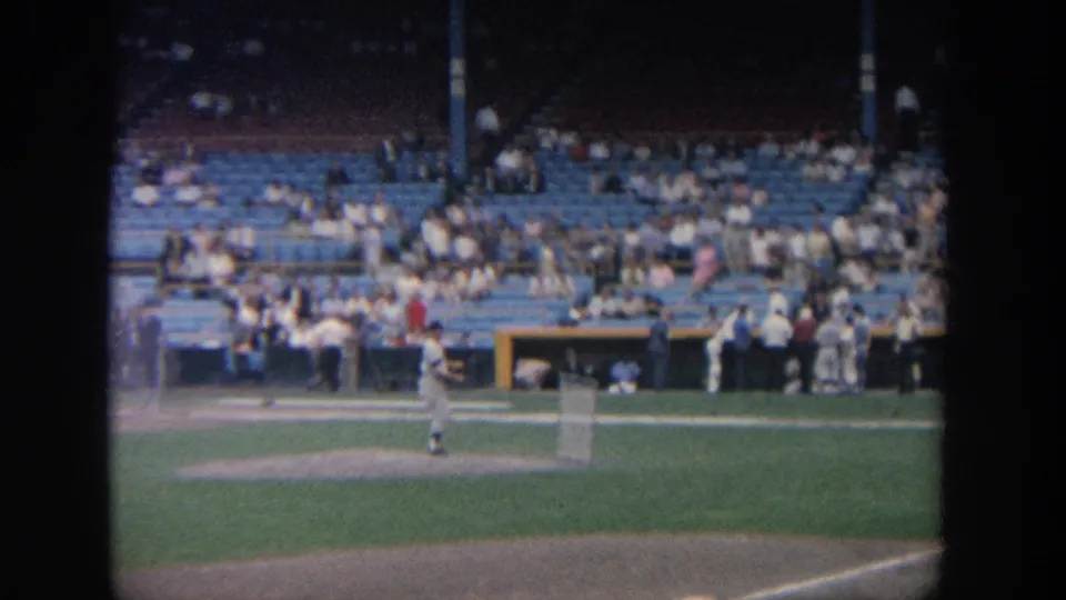 1964 New York 8mm frame showing baseball players warming up near the infield as spectators sit in the stands.