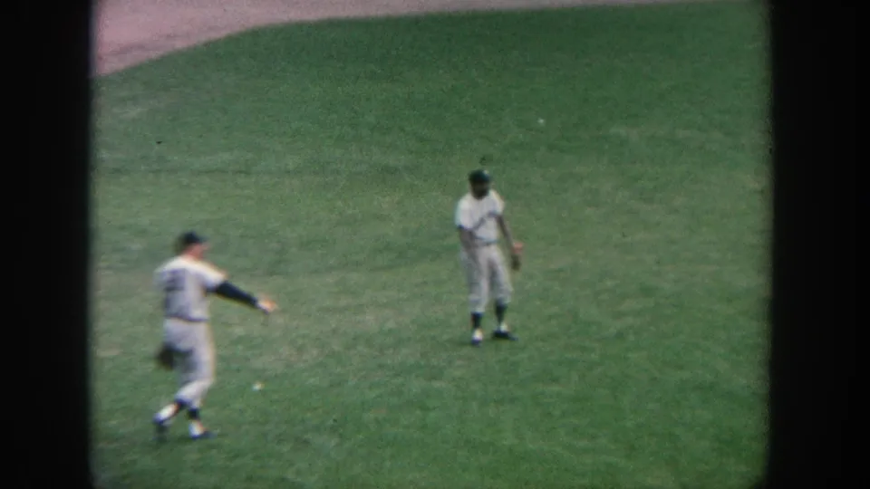 1964 New York 8mm footage shows two baseball players on green outfield grass, with one moving at the left edge of the frame.