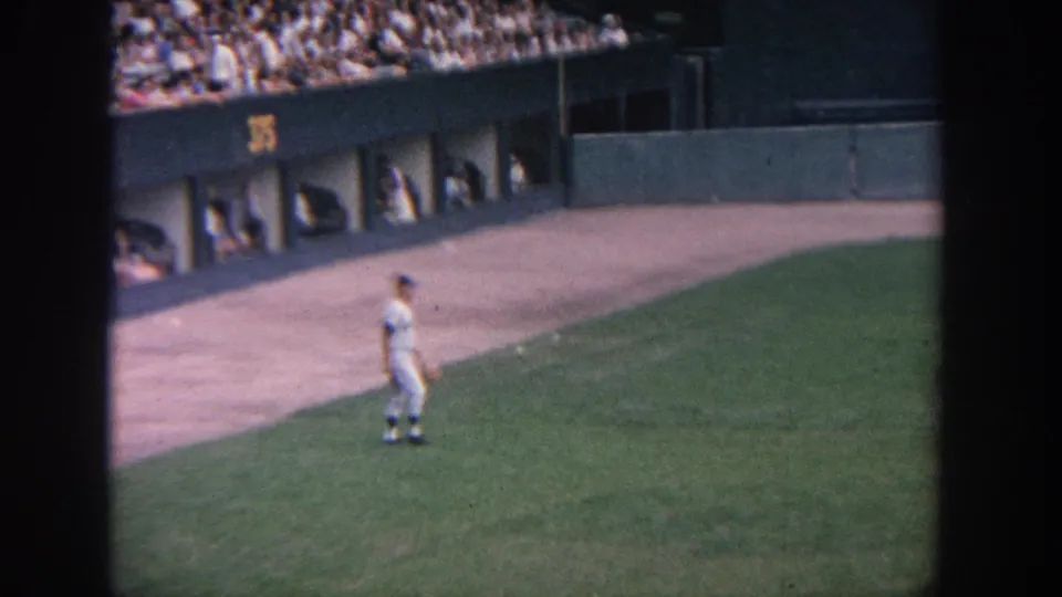 1964 New York 8mm footage shows a uniformed baseball player near the warning track, stadium wall, and seated crowd.