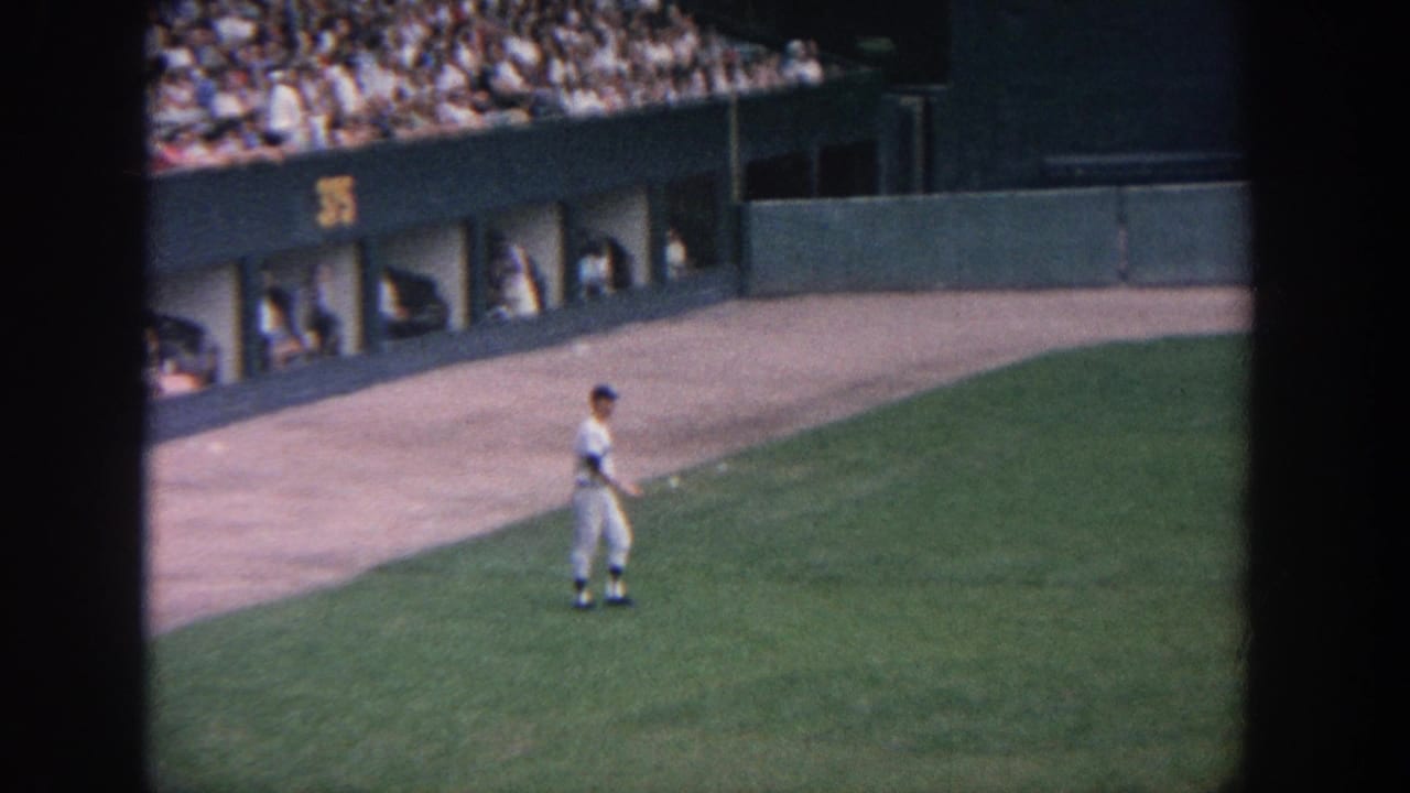 1964 New York home-movie frame showing a baseball player on the grass near the dugout area, with spectators above.