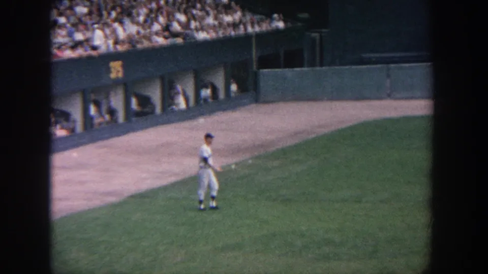 1964 New York home-movie frame showing a baseball player on the grass near the dugout area, with spectators above.