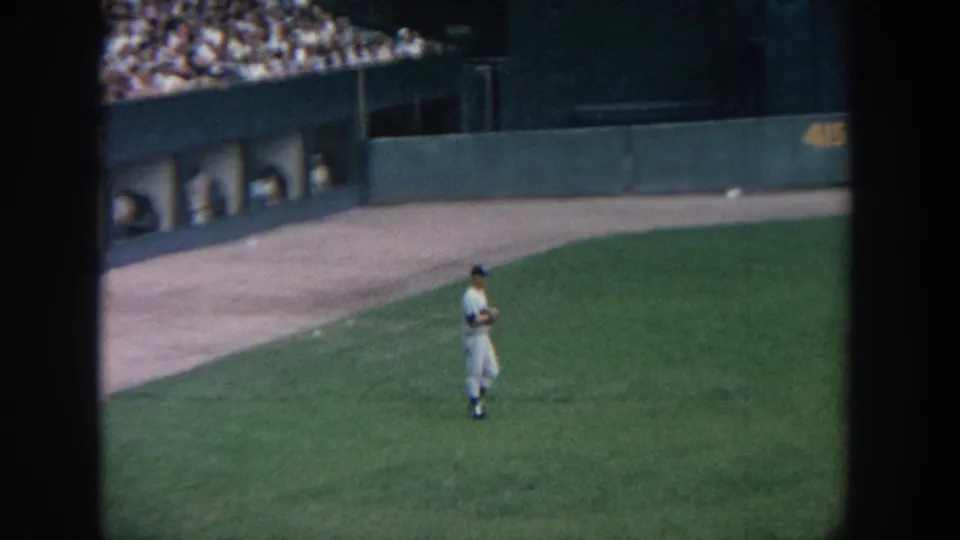 Home-movie view of a baseball player paused on the field, with dugout bays and spectators visible in the background.
