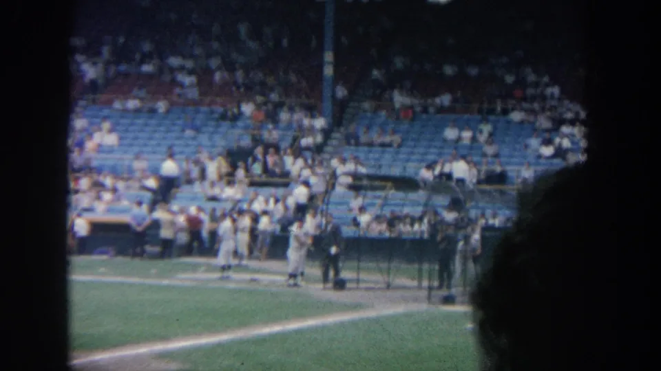 1964 New York baseball stadium frame showing players near the baseline and stands with scattered spectators.