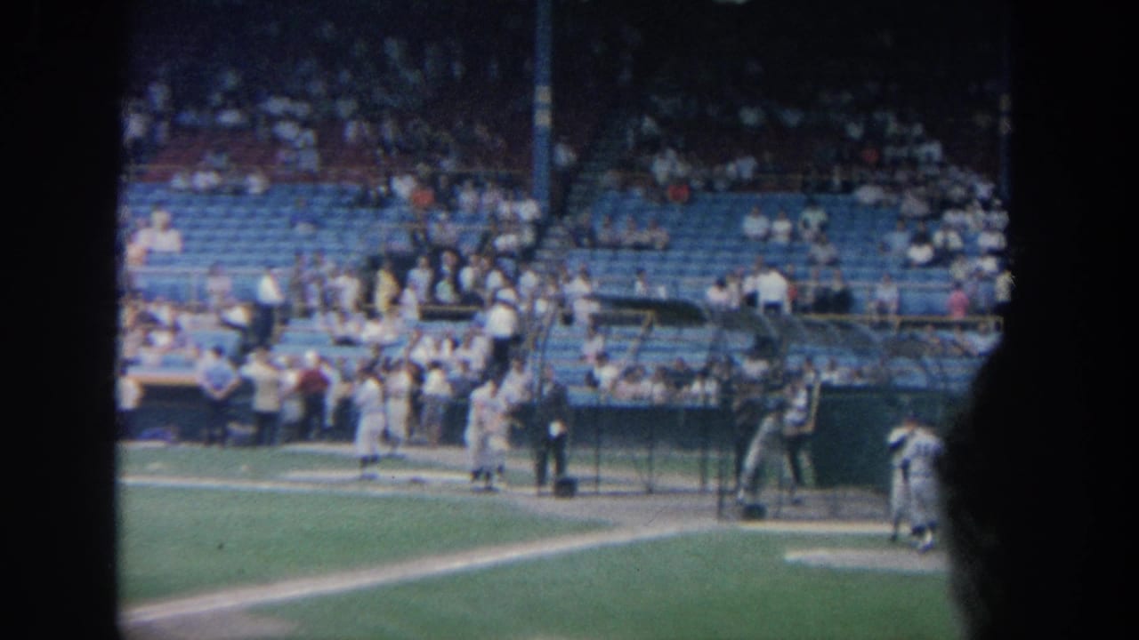 1964 New York 8mm footage shows baseball players in white uniforms near batting cages with spectators visible in blue stadium seating.
