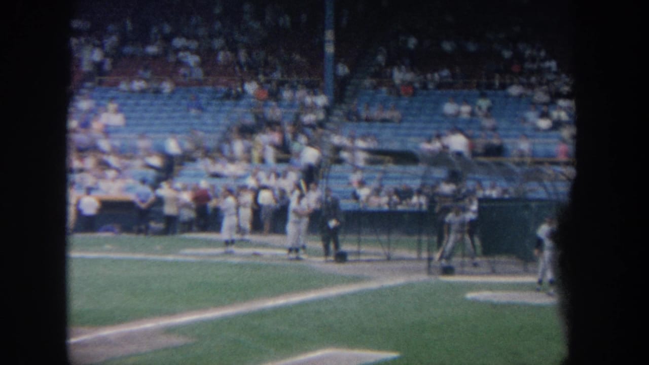 A batter stands near the plate while players and spectators are visible around the batting cage area.