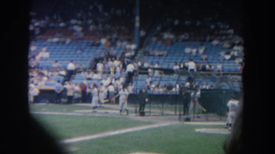 1964 New York 8mm frame shows several baseball players around protective cages on the field, with a crowd scattered across blue stadium seating.