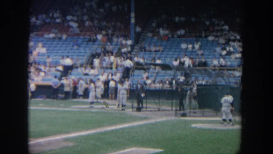 1964 New York stadium footage shows a player at a protective batting cage, teammates nearby, and spectators seated beyond the field.