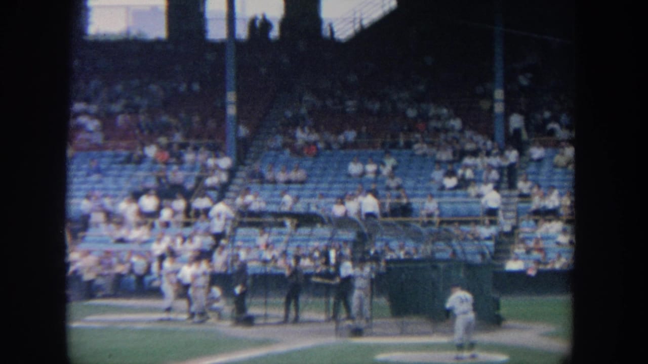 1964 New York 8mm view of baseball players preparing near home plate with blue stadium seats and spectators behind them.