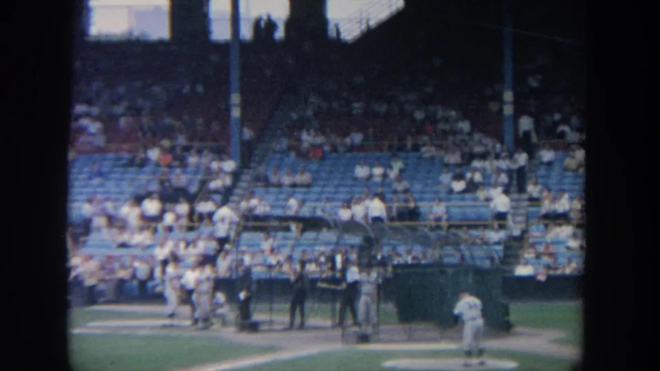 1964 New York 8mm view of baseball players preparing near home plate with blue stadium seats and spectators behind them.