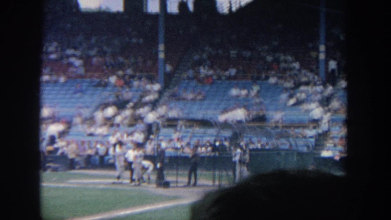 Baseball players near the dugout and home plate with fans in the stands.