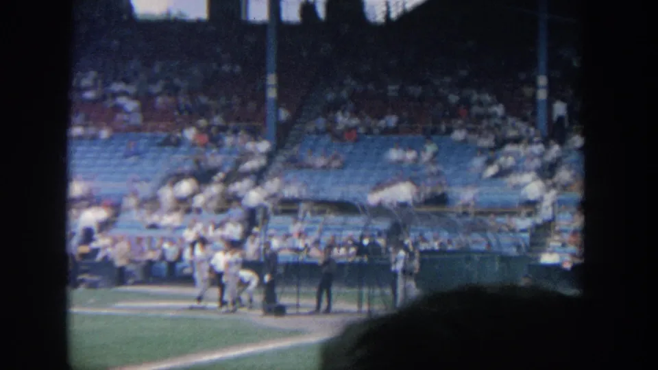 1964 New York baseball stadium scene with players clustered near the dugout and spectators seated behind the field.