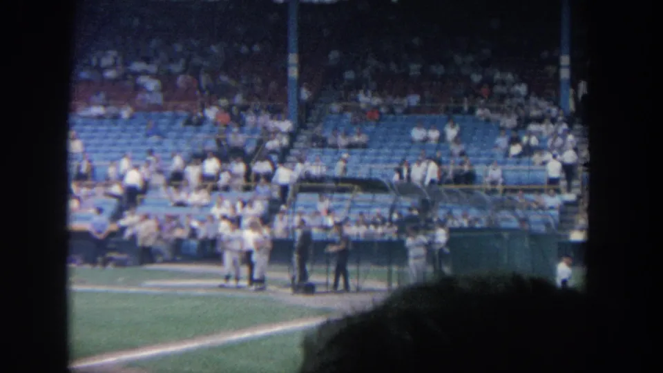 1964 New York 8mm stadium view showing players and field personnel near home plate with a seated crowd behind them.