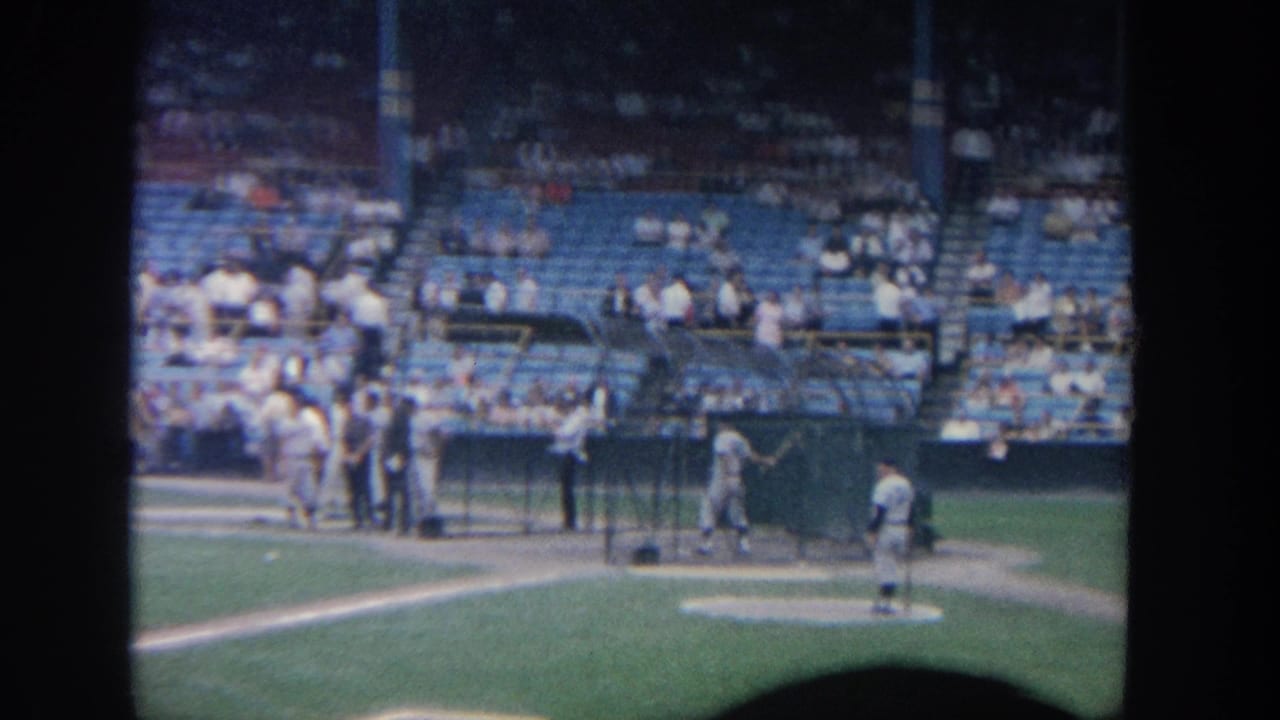 1964 New York home-movie frame showing players near a batting cage, a pitcher on the mound, blue stadium seats, and spectators in the background.